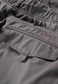 Gros plan sur un pantalon en tissu gris avec une ceinture élastique et une poche à rabat sécurisée par une petite languette en tissu.