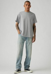 Camiseta de algodón gris con mangas cortas, combinada con pantalones vaqueros de mezclilla azul claro. El modelo lleva zapatillas negras y blancas. Ajuste y diseño casual.