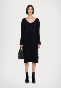 Rochie lungă neagră tricotată cu decolteu rotund. Modelul ține o geantă neagră, purtând șosete negre până la gleznă și pantofi cu vârf ascuțit.