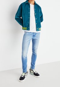 Persona con chaqueta de botones a la altura del pecho en color verde azulado, camiseta blanca, jeans ajustados de color azul claro y zapatillas Nike en blanco y negro, de pie sobre un suelo blanco.