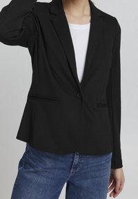 Černý blazer vyrobený z měkké tkaniny, s klopovým límcem, jedním knoflíkem a dvěma předními kapsami. Nosený přes bílou košili.