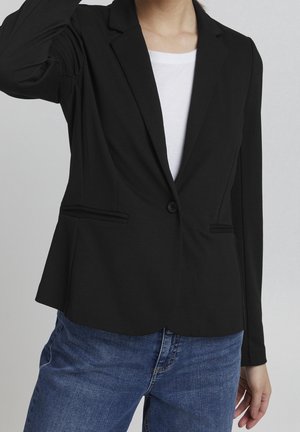 Schwarzer Blazer aus weichem Stoff, mit Reverskragen, Ein-Knopf-Verschluss und zwei vorderen Taschen. Über einem weißen Hemd getragen.
