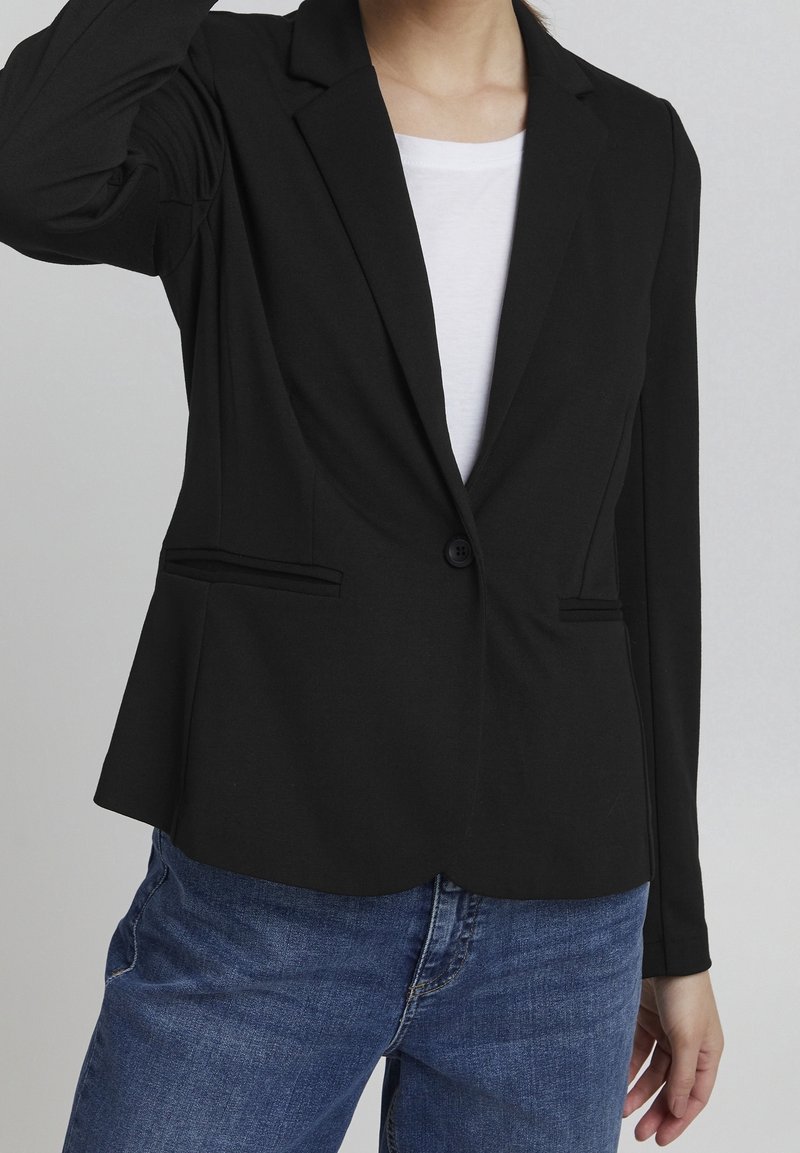 Černý blazer vyrobený z měkké tkaniny, s klopovým límcem, jedním knoflíkem a dvěma předními kapsami. Nosený přes bílou košili.