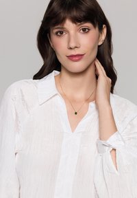 Złoty naszyjnik z zielonym wisiorkiem noszony na szyi modelki, dopełniający białą, fakturowaną bluzkę z podwiniętymi rękawami.