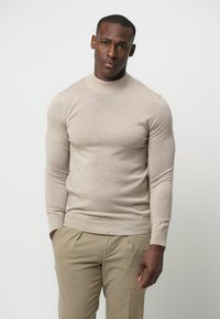 Beige gebreide sweater met een hoge halslijn, lange mouwen en een slim fit. Gecombineerd met lichtgekleurde broeken, met een gladde textuur overal.