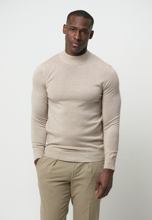 Beige Strickpullover mit hohem Kragen, langen Ärmeln und schmaler Passform. Kombiniert mit hellen Hosen, durchgehend glatte Textur.