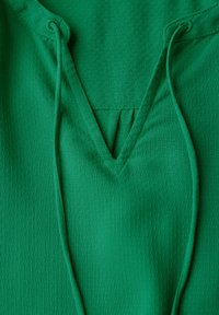 Top sportivo verde con tessuto texturizzato, scollatura a V e dettagli con cordini. Il materiale sembra leggero e traspirante.