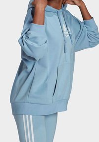 Ljusblå oversized hoodie i mjukt tyg med kanguruficka, dragsko i huvan och vita logotypdetaljer på bröstet.