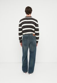 Prúhovaný sveter v tmavohnedej, krémovej a šedej farbe, skombinovaný s modrými, voľne strihanými džínsami s dvoma zadnými vreckami a hnedou koženou nášivkou.