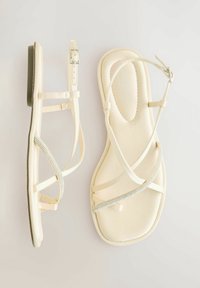 Sandali piatti di un colore crema chiaro, con cinghie sottili e dettagli in strass. La suola è liscia con una leggera curvatura. Dettaglio della fibbia in argento.