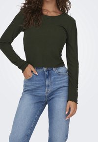 Langärmeliges, olivgrünes, geripptes Oberteil mit rundem Ausschnitt und gewelltem Saum, kombiniert mit hellblauen, hoch taillierten Jeans.