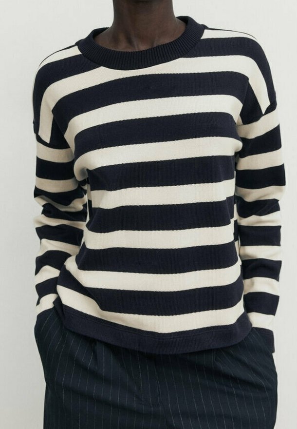 Personne portant un pull à rayures horizontales noires et blanches, les mains dans les poches d'un pantalon sombre à fines rayures.