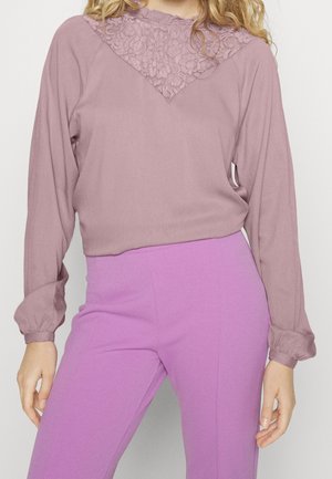 Langærmet top i afdæmpet mauve med blonde detaljer på en v-formet udskæring, parret med højtaljede lilla bukser, blød stoftekstur.
