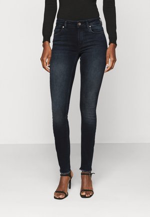 Jeans skinny azul escuro com uma textura suave, cintura média, e desbotamento subtil, combinados com sapatos de salto pretos com tiras.