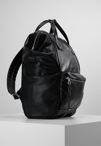 Mochila de cuero negro con una textura suave, que cuenta con dos asas en la parte superior, correas de hombro ajustables y un bolsillo frontal con cremallera.