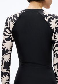 Žmogaus nugaros vaizdas, dėvinčio juodą ilgomis rankovėmis viršutinius drabužius su smėlio spalvos gėlėtais raštais ant rankų ir pečių.