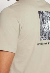 Beige tričko s černobílou grafikou a textem "THE NORTH FACE" a částečně viditelným "NEVER STOP EXPLORING" na zadní straně.