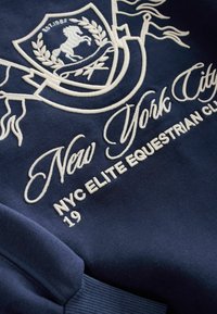 Navy sweatshirt featuring embroidered crest with a horse, "New York City" and "NYC Elite Equestrian Club" text in white, textured fabric.