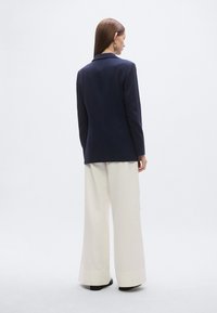 Marynarka marynarska w kolorze granatowym z wyciętymi klapami, dopasowany krój, zestawiona z kremowymi spodniami o szerokich nogawkach. Tkanina wydaje się gładka, z delikatnym połyskiem i czystymi liniami.