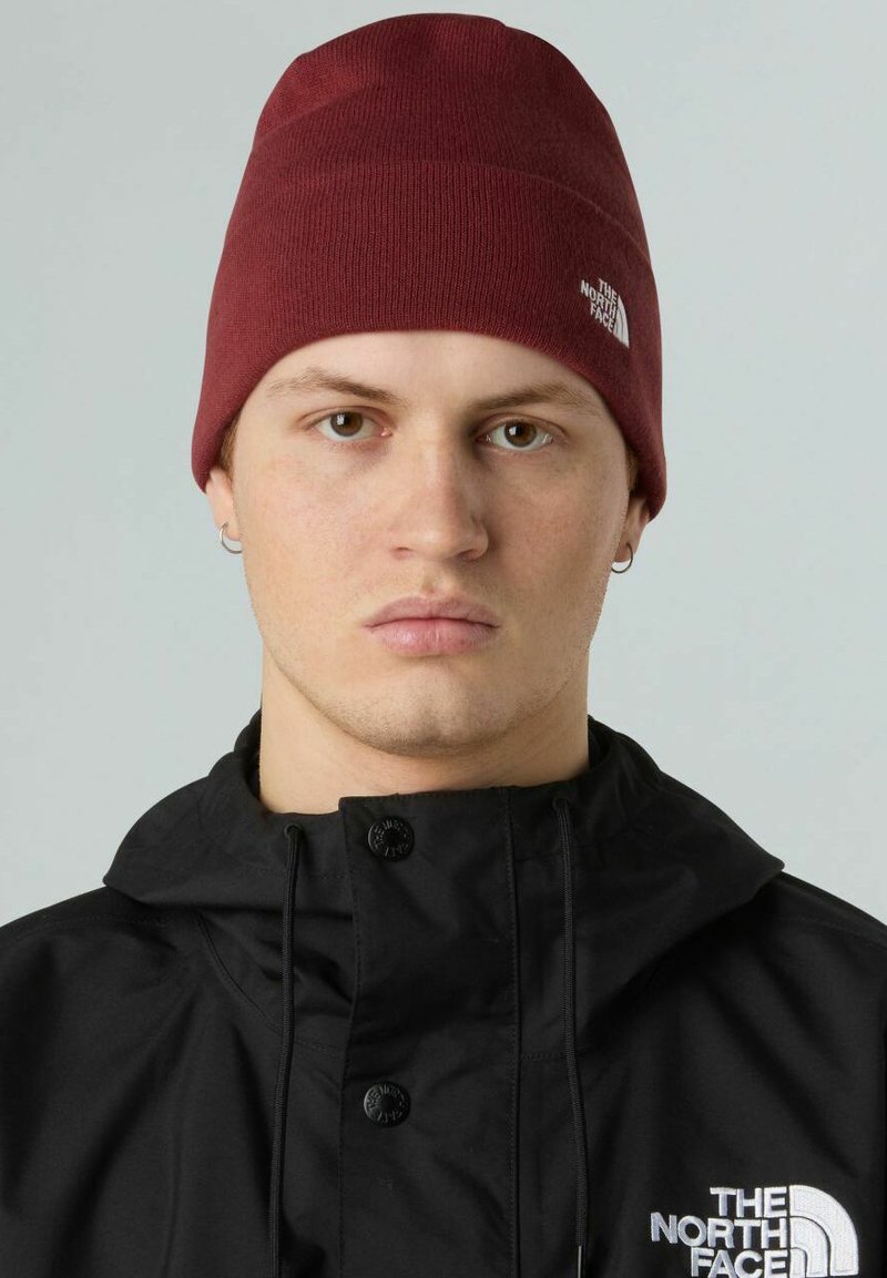 Beanie lavorato a maglia borgogna con una vestibilità aderente, caratterizzato da sottili righe orizzontali e un piccolo dettaglio del logo bianco. Indossato con una giacca nera.