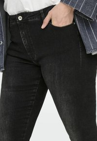 Melni denim džinsi ar šauru piegriezumu, redzamu priekšējo kabatu un metāla pogu aizdari. Audumam ir neliela tekstūra un izbalējis izskats.