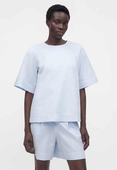 Γυναίκα με σκουρόχρωμο δέρμα που φοράει γαλάζιο υφασμάτινο κοντομάνικο μπλουζάκι και ταιριαστά σορτς, στέκεται σε απλό φωτεινό φόντο.