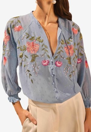 Vrouw draagt een lichtblauw blouse met roze en groene bloemenborduursels, knopen bedekt met stof, en beige broek, hand in de zak.