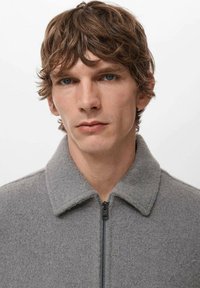 Giacca grigia in lana con collo e cerniera. Superficie testurizzata. Il modello ha capelli corti e ondulati e guarda verso la fotocamera su uno sfondo neutro.