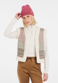 Fejkpälsväst med mönstrad stickad framdel i pastellfärger, kombinerad med en rosa mössa. Vit långärmad topp och bruna byxor fulländar looken.