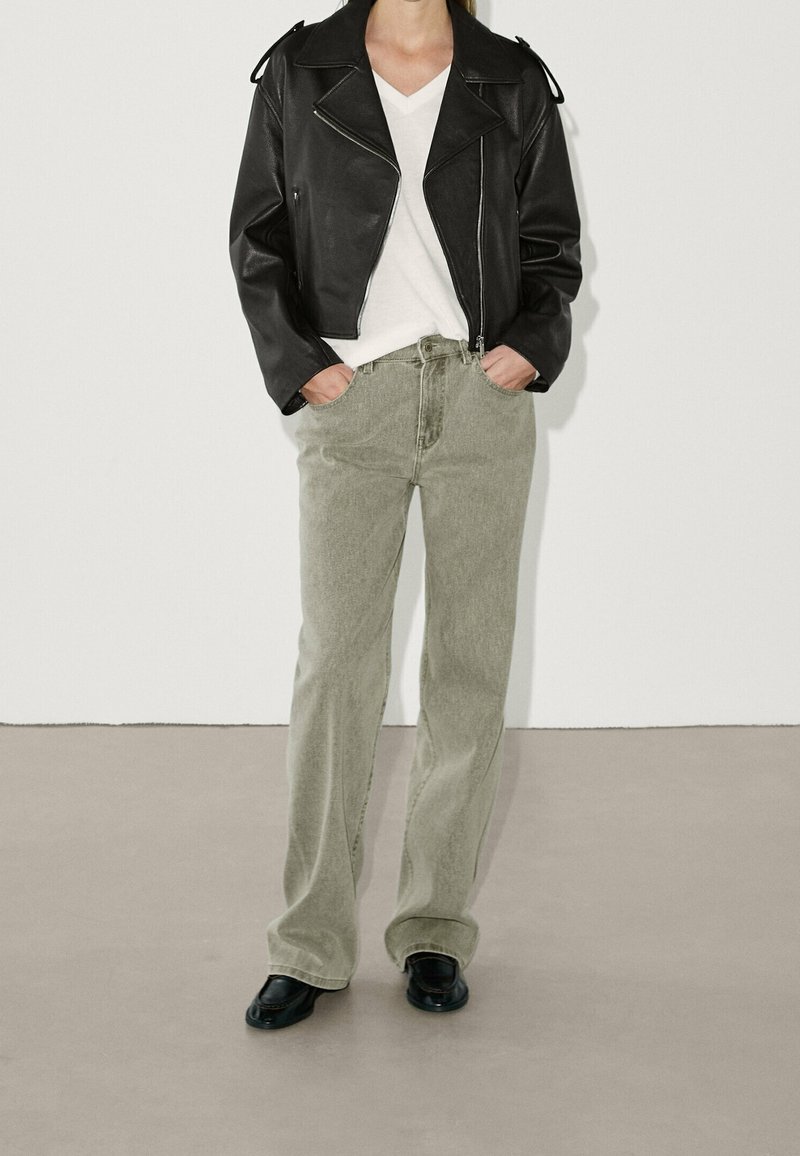 Svart läderbikerjacka, vit V-ringad t-shirt, ljusgröna raka jeans och svarta skor med rund tå. Minimalistisk design.