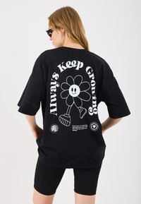 T-shirt oversized preta com um gráfico de flor branca e o texto "Sempre Continua a Crescer" nas costas. Manga curta e ajuste relaxado.