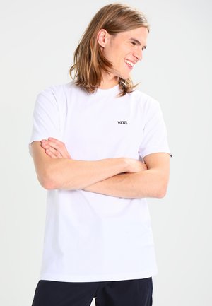 Camiseta blanca de manga corta hecha de algodón, con un pequeño logo negro de "VANS" en el pecho, combinada con pantalones oscuros. Textura suave.