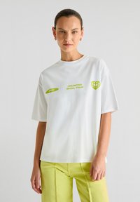 Witte katoenen t-shirt met oversized pasvorm, met groene grafische tekst en vormen op de voorkant. Gepaard met lichtgroene broek.
