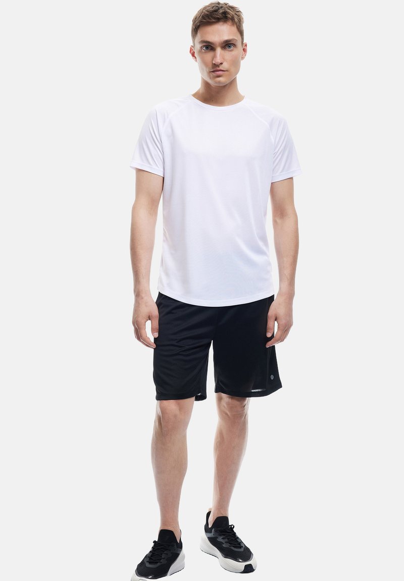 Hvid kortærmet træningsskjorte og sorte shorts. Skjorten har en rund hals; shortsene er knælange med en glat tekstur. Sorte sneakers fuldender outfittet.