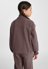 Brun fleecejacka med hög krage, mjuk textur och avslappnad passform, bärs över matchande byxor, vy från baksidan.