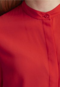 Camicia rossa con bottoni, caratterizzata da un colletto arrotondato, una texture liscia e una patta nascosta con chiusura a un solo bottone al collo.
