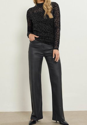 Femme portant un haut noir transparent à manches longues à imprimé léopard, un pantalon en cuir noir taille haute et des talons noirs à bouts pointus, debout contre un mur blanc.