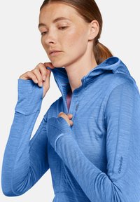 Sudadera azul claro con cremallera en un material suave y texturizado; cuenta con capucha, agujeros para los pulgares y un logo sutil en la manga.