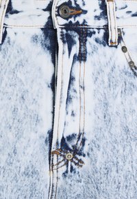 Pantalones vaqueros de mezclilla con un patrón tie-dye en azul claro y blanco, con costuras naranjas contrastantes y botones de metal plateado en la cintura.