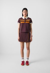 Mlada žena nosi vino crvenu Adidas polo košulju i odgovarajuću suknju s narančastim trakama, bijele čarape s crnim trakama, crvene tenisice i narukvicu.