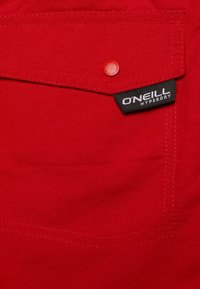 Tasca in tessuto rosso con un flap arrotondato, fissato da un bottone abbinato. Presenta un'etichetta nera con testo "O'Neill Hyperdry". Dettagli di cuciture testurizzate.