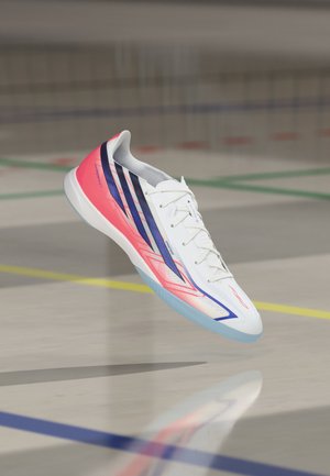 Weißer und pinker Hallensportschuh mit blauen Akzenten und Schnürsenkeln, der über einem Gymboden mit farbigen Linien schwebt.