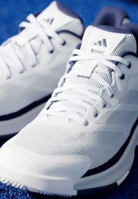 Bílé sportovní boty s texturovaným svrškem, šněrováním a tmavě modrým akcentem. Mají výrazné logo a gumovou podrážku pro lepší přilnavost.