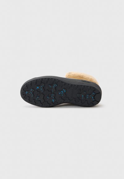 Suela de una bota con una superficie de goma negra texturizada que presenta un patrón de banda de rodadura prominente. Parte superior de gamuza de color marrón claro visible en el borde superior.