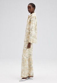 Beige jas met bloemenpatroon en wijde broek in zachte stof. De outfit heeft een kraag en een relaxte pasvorm. Zilveren hakken accentueren de look.