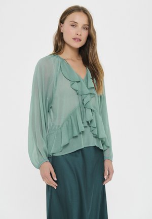 Ljusgrön blus med långa, puffiga ärmar, prydd med lager av volanger längs halsringningen och nederkanten. Mjukt, genomskinligt tyg med en svepande design.