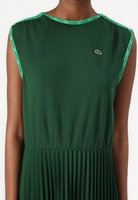 Zelena plisirana obleka z okroglim izrezom, zaključena z zelenim Lacoste trakcem s potiskom ob ramenih in majhnim Lacoste logotipom na prsih.