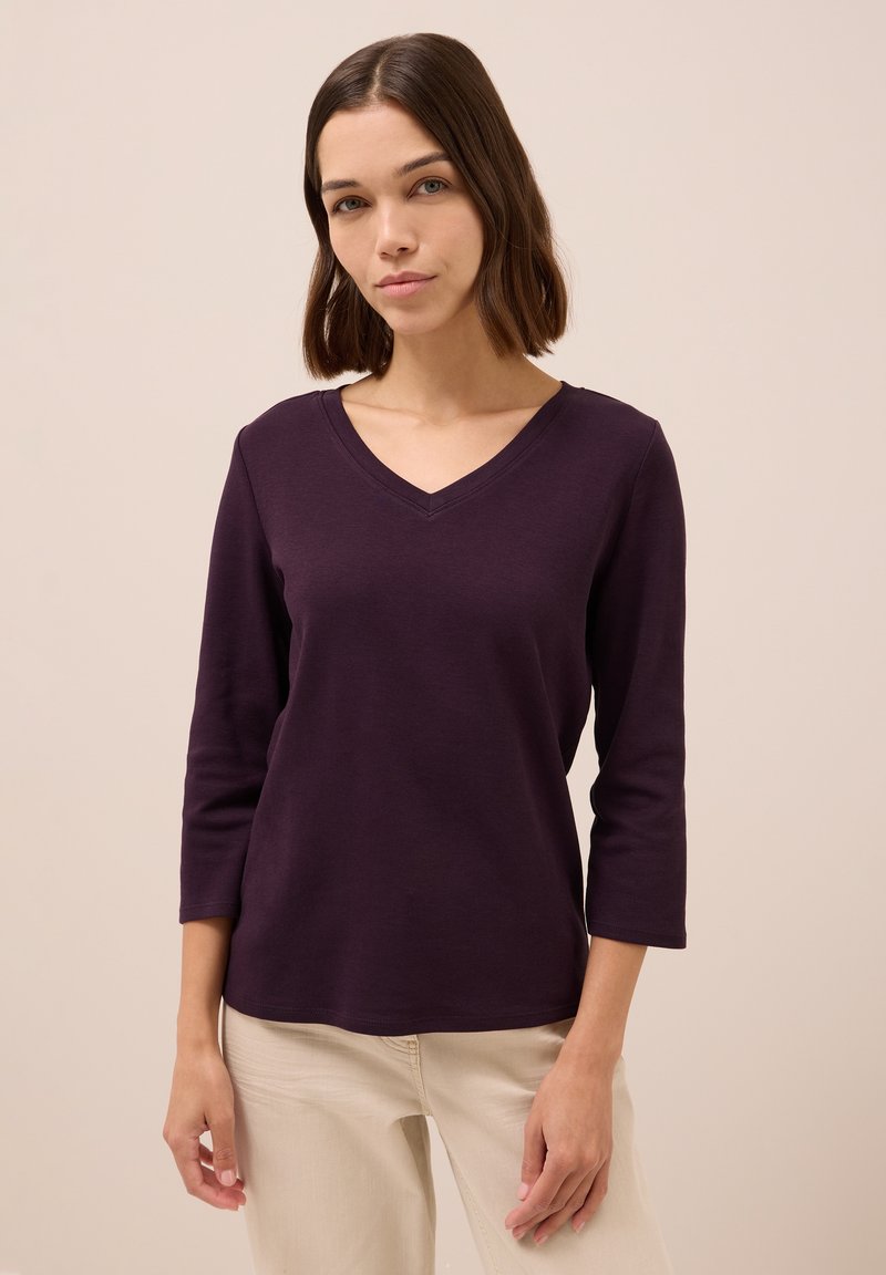 Top a manica lunga con scollatura a V in tessuto viola scuro. Texture morbida con vestibilità rilassata. Indossato con pantaloni chiari, dettagli minimi.
