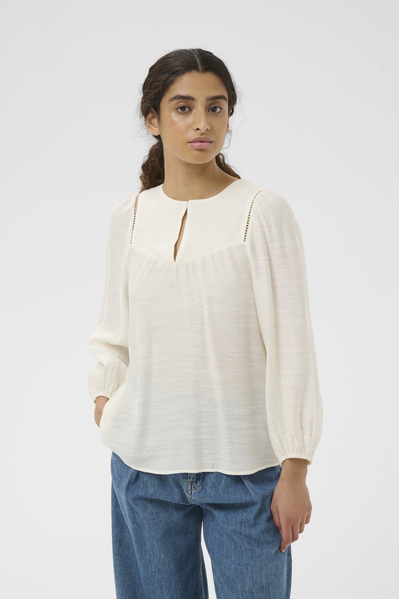 Femme aux cheveux foncés tirés en arrière, portant un chemisier blanc ample à encolure ajourée et un jean bleu, debout devant un fond uni.