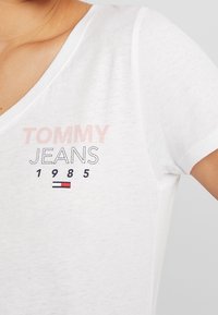 Camiseta blanca con escote en V, que presenta el texto "TOMMY JEANS" en rosa claro y negro, con el año "1985" y un acento de logo en rojo/azul.